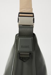 Borsa impermeabile verde scuro con tracolla regolabile beige e fibbia in metallo argentato, dotata di una toppa ricamata con l'etichetta "RAINS."