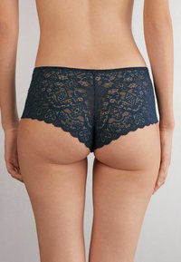 Granatowe koronkowe majtki hipster nawiązujące do kwiatowych wzorów, z falbaniastymi brzegami i półprzezroczystą fakturą, oferujące dopasowany krój i pełne pokrycie.