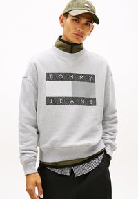 Γκρι φούτερ με ψηλό γιακά, με αντίθετο γραφικό "TOMMY JEANS" σε μαύρο και γκρι. Φορεμένο πάνω από μια καρό πουκάμισο.