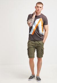 Camiseta gráfica marrón con rayas naranjas y azules, combinada con pantalones cortos de carga en verde oliva y zapatillas grises. El modelo tiene tatuajes.