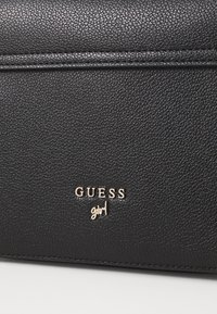 Črna teksturirana torba iz umetnega usnja s zlatim logotipom "GUESS" in besedo "girl" v dodelanem fontu. Čist dizajn z diskretnimi šivi.