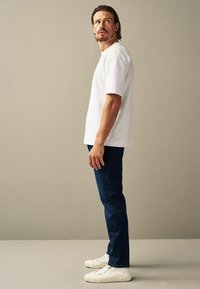 Tricou alb din bumbac cu mâneci scurte și o croială lejeră, purtat cu blugi albaștri închisi și adidași albi. Stând pe un fundal neutru.