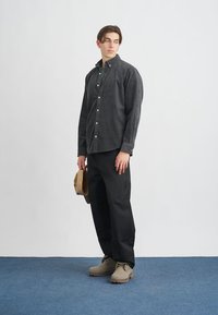 Γκρι καμπαρντίνα πουκάμισο με κουμπιά, μαύρα παντελόνια και μπεζ παπούτσια. Κρατώντας ένα καφέ καπέλο, στέκεται σε μπλε χαλί μπροστά σε λευκό φόντο.