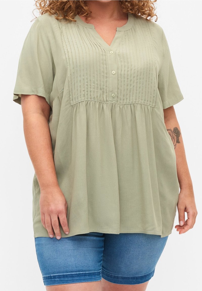 Light green blouse with short sleeves, button placket, and pleated detailing at the chest. Paired with denim shorts. Soft fabric texture.