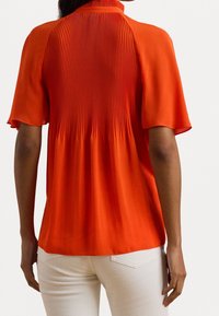 Orange silk blouse featuring pleated detailing at the back and short sleeves, paired with light-colored pants. Smooth texture and relaxed fit.