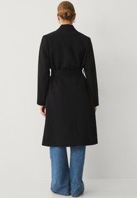 Sort lang jakke med bælte, notch krave og lange ærmer, parret med blå bredbenede jeans. Glat tekstur, minimalistisk design.
