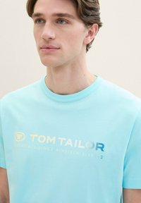 Světle modré bavlněné tričko s kulatým výstřihem; na přední straně je potisk "TOM TAILOR" ve gradientu od bílé po modrou; krátké rukávy, hladká textura.