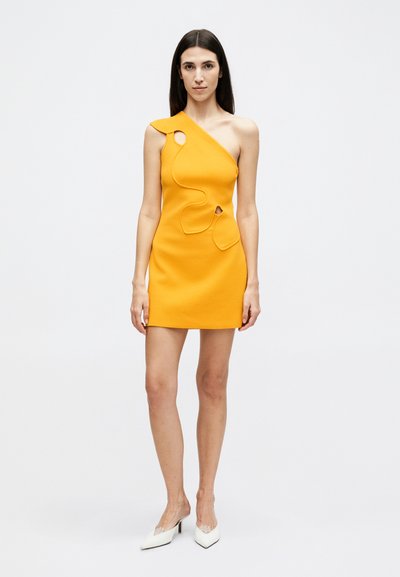 Femme aux longs cheveux foncés portant une mini robe jaune asymétrique sans manches avec des détails découpés, debout devant un fond blanc uni.