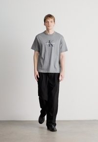 Γκρί βαμβακερό μπλουζάκι με λογότυπο "Calvin Klein", συνδυασμένο με μαύρο παντελόνι και μαύρα παπούτσια. Μοντέλο που περπατά σε λείο δάπεδο με απλό φόντο.