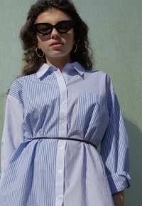 Modrá a bílá pruhovaná košile s dlouhými rukávy, s knoflíkovým zapínáním a černým páskem. Modelka nosí černé sluneční brýle ve tvaru kočičích očí a kruhové náušnice.