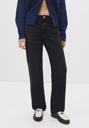 Kobieta ubrana w czarne, wysoki stan jeansy, granatowy, krótki kardigan, złote pierścionki i czarno-białe loafersy, stojąca na białym tle.