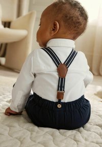 Temno modre kratke hlače z elastičnim pasom in naramnicami; bela srajca s ovratnikom; rjav usnjen pas z gumbom; teksturirana tkanina.