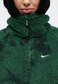 Πράσινο φλις τζάκετ με ψηλό γιακά, υφασμάτινη υφή, φερμουάρ κλεισίματος και λευκό λογότυπο Nike. Το υλικό έχει πλούσια, άνετη όψη.