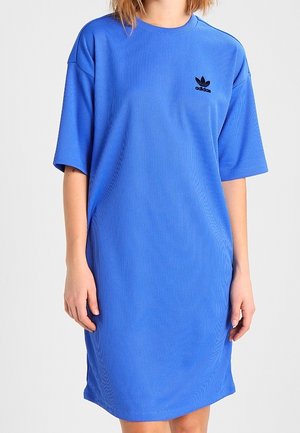 Vrouw die een loszittende, knielange blauwe Adidas-jurk draagt met korte mouwen en een zwart logo op de borst.