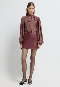 Bordowa bluzka z bufiastymi rękawami i detalem w postaci kokardy, w zestawie z dopasowaną burgundową skórzaną spódnicą z złotymi zamkami błyskawicznymi. Czarne buty na odkrytej pięcie.