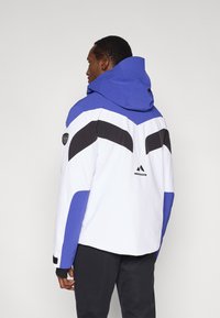 Bunda s farebnými blokmi v bielej, modrej a čiernej farbe. Má priestrannú kapucu, uhlové dizajny a logo patch na rukáve. Textúrovaná tkanina.