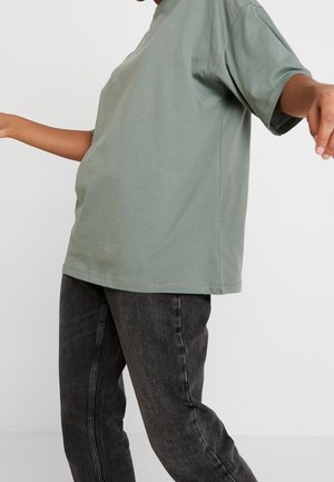 Person wearing a loose sage green short-sleeve shirt and faded black jeans against a plain light gray background.