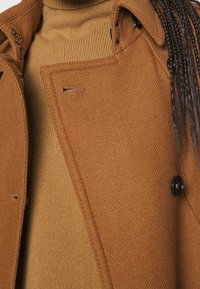 Brązowy płaszcz z mieszanki wełny o teksturowanym wykończeniu i dużymi czarnymi guzikami, nałożony na beżowy prążkowany golf.