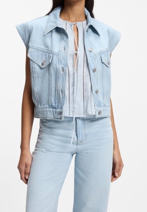 Kvinde iført lyseblå ærmeløs denimjakke over hvid bluse med bindebånd foran og matchende lyseblå denimjeans.