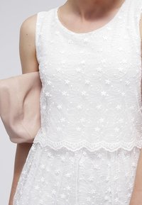 Weißes ärmelloses Kleid mit floralen Stickereien und wirbelnden Mustern. Der Rock ist durchsichtig und über einer soliden Unterschicht geschichtet, mit einem gewellten Saum.