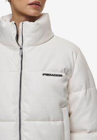 Hvit polstret jakke med høy krage, med svart glidelås og logo på brystet. Glatt tekstur, moderne design og normal passform.