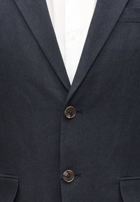 Κοντινή λήψη ενός σκούρου γκρι σακακιού με δύο κουμπιά κουμπωμένα πάνω από ένα λευκό πουκάμισο.