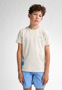 T-shirt de algodón beige claro con estampados gráficos azules, con cuello redondo y mangas cortas, combinado con pantalones cortos azul claro.