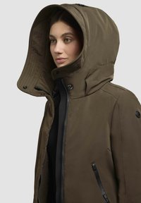 Veste à capuche vert olive avec fermeture éclair, grande capuche doublée et poches latérales, dotée d'accents en métal noir et d'une texture lisse.