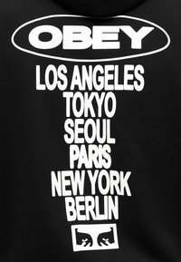 Μαύρο φούτερ με μεγάλο λευκό κείμενο. Στο μπροστινό μέρος εμφανίζεται το "OBEY" και τα ονόματα πόλεων: Λος Άντζελες, Τόκιο, Σεούλ, Παρίσι, Νέα Υόρκη, Βερολίνο, με ένα γραφικό από κάτω.