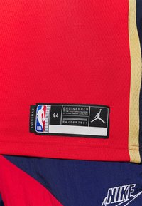 Red basketball jersey with mesh texture, featuring a black label with NBA logo, size 44, and Jordan branding. Navy and gold accents included.