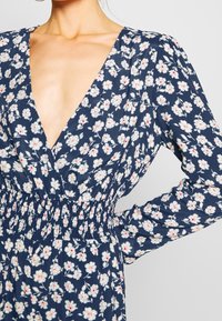Mörkblå blommig klänning med djup V-ringning, långa ärmar och smockad midja. Har vita blommor med röda detaljer på ett texturerat tyg.