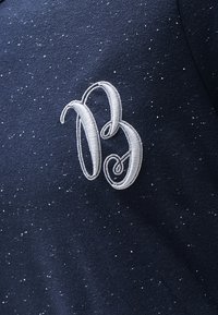 Námořnická modrá látka se skvrnitou texturou s bílou vyšívanou literou "B" umístěnou na levé straně.