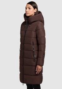 Manteau rembourré marron avec un col haut et une grande capuche, doté de poches latérales zippées et de fermetures éclairs noires contrastantes à l'avant et sur les manches.