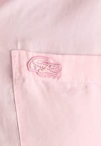 Tessuto di cotone rosa con texture diagonale; presenta un piccolo logo ricamato a forma di coccodrillo di un rosa più chiaro sulla tasca del petto.