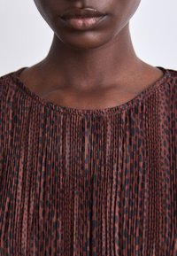 Haut plissé marron avec un col rond, présentant un motif de taches sombres et de rayures, fabriqué en tissu léger.