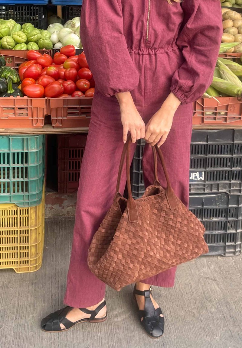 Persona con mono rojo sosteniendo un gran bolso marrón de mimbre, de pie cerca de cajas con verduras frescas que incluyen tomates, calabacines y patatas.