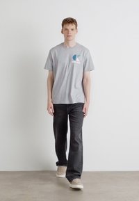 Grå bomulds t-shirt med et blåt og hvidt logo, parret med mørk denim jeans og beige sneakers, præsenteret mod en lys baggrund.