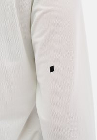 White athletic long-sleeve shirt featuring a textured, breathable fabric with a small black square logo on the sleeve.