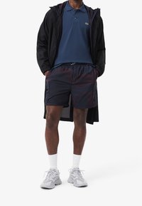 Veste légère noire, polo bleu marine, shorts bleu marine avec poches latérales, et baskets grises avec chaussettes blanches, se tenant devant un fond blanc.