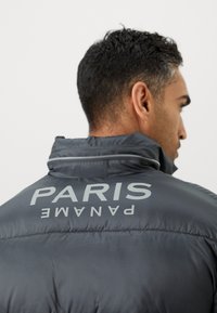 Siva puf jakna s visokim ovratnikom i velikim natpisom "PARIZ PANAMA" na leđima. Materijal ima glatku, sjajnu teksturu.