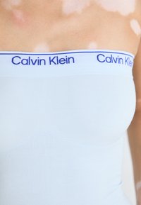 Svijetloplavi bez naramenica s gornjim dijelom s glatkom teksturom i kontrastnim bijelim pojasom s natpisom 'Calvin Klein' u plavom tekstu duž gornjeg ruba.