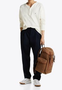Cream long-sleeve henley shirt, navy pleated pants, white sneakers with brown accents, and a brown fabric backpack with red hardware details.