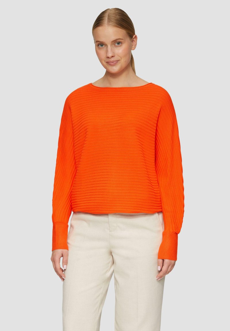 Oranger gerippter Pullover mit weitem Ausschnitt und langen Ärmeln. Kombiniert mit cremefarbenen Hosen. Strukturiertes Stoff sorgt für Dimension.