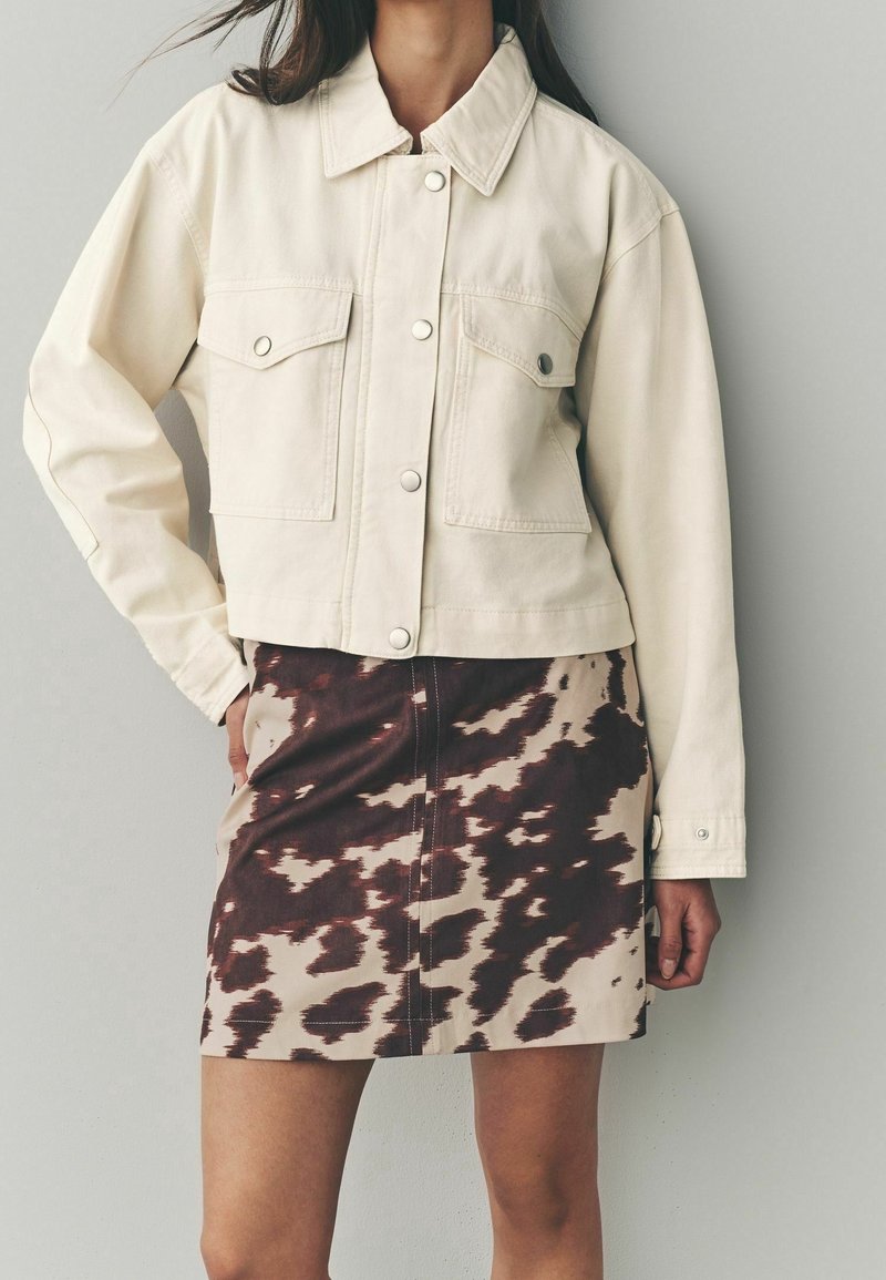 Femme portant une veste courte crème avec boutons-pression et grandes poches avant, associée à une mini-jupe à motif abstrait marron et beige.
