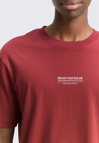 Zbliżenie na osobę w czerwonej koszulce z białym napisem "DENIM TOM TAILOR" oraz mniejszym hasłem na lewym zakresie klatki piersiowej.