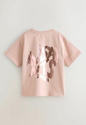 Svijetlo ružičasta majica s kratkim rukavima s sjajnim, iskrivljenim tekstom "ALSANTS LONDON" na leđima, prikazana na jednoličnoj svijetloj pozadini.