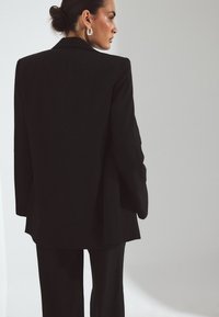 Črn tailored blazer in usklajene hlače, ki nudita elegantne linije in minimalističen dizajn, z teksturirano tkanino in elegantnimi klapnama.