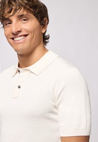 Polo shirt beige a maniche corte realizzato in morbido tessuto, con colletto rilassato e polo a due bottoni con dettaglio del bottone nero.