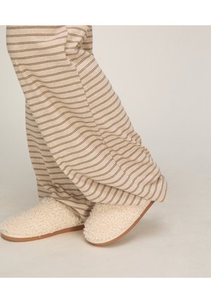 Persoon die beige gestreepte broek en crèmekleurige pluizige pantoffels draagt, staat op een lichte vloer met één voet iets opgeheven.