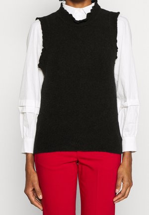 Personne portant un gilet en maille noir sans manches sur une chemise blanche à manches longues avec des poignets volantés et un pantalon rouge.